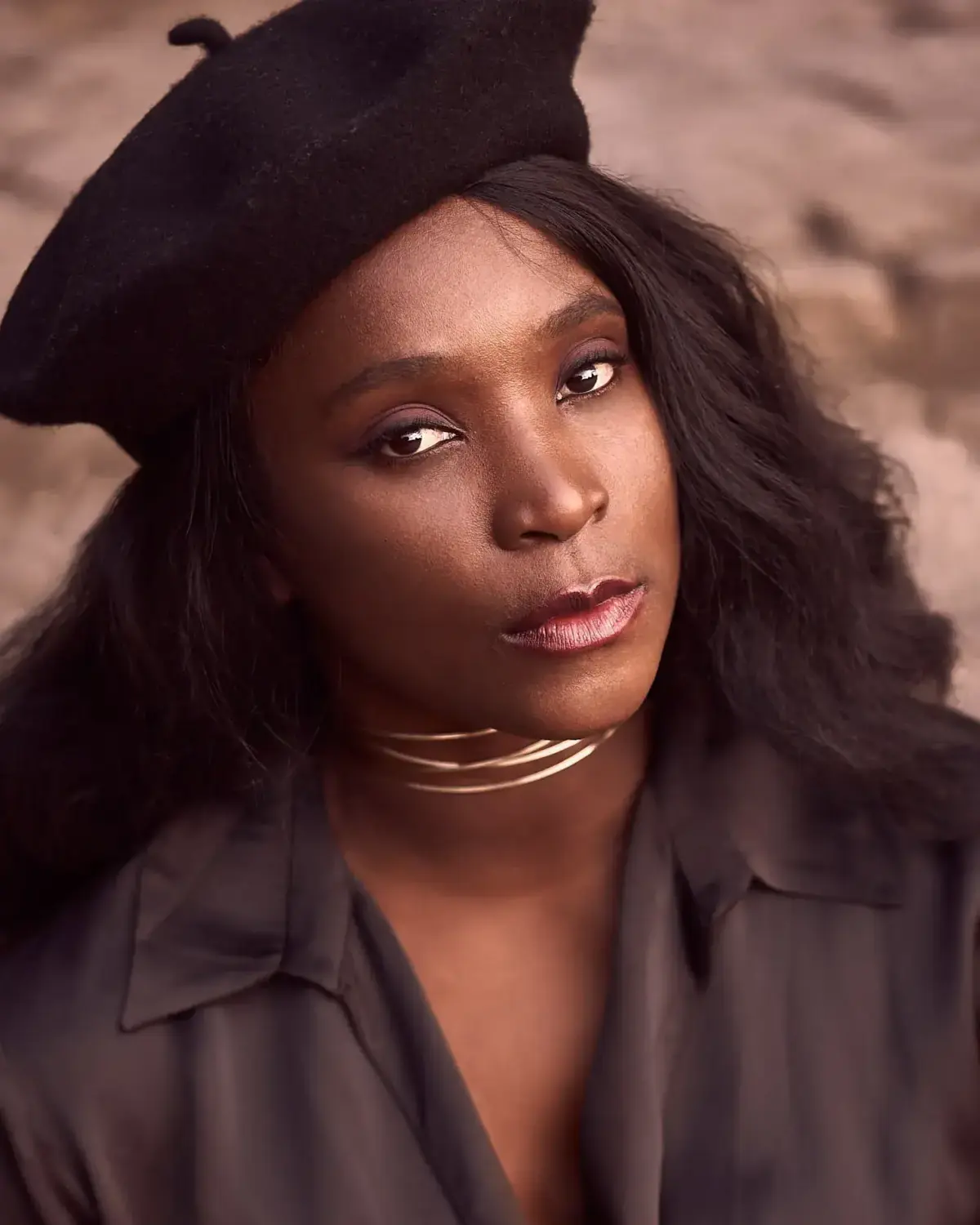 Nahaufnahme einer Frau mit schwarzer Baskenmütze und offenem dunklem Haar; dezentes Make-up, goldener Halsreif in drei Ringen, ruhiger Blick; weiches Außenlicht auf neutralem Hintergrund.