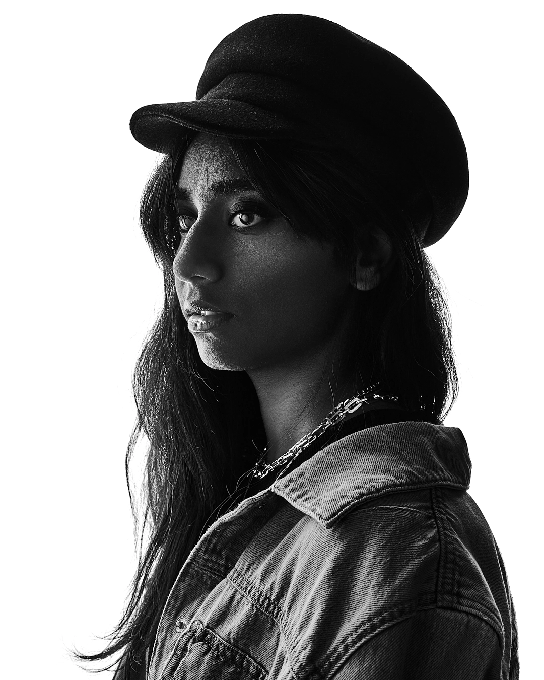 Schwarz-Weiß-Profilaufnahme einer Frau mit dunkler Schirmmütze und langen Haaren; strukturierte Jeansjacke, klare Lichtkante; neutraler, heller Hintergrund mit starkem Kontrast.