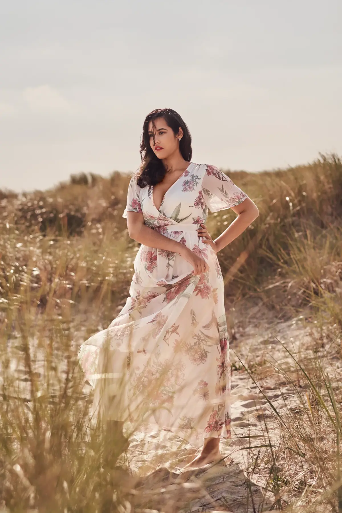 Eine Frau steht am Strand zwischen Dünen und Gras. Sie trägt ein langes, leicht wehendes Kleid mit Blumenmuster und hat eine Hand in der Hüfte.