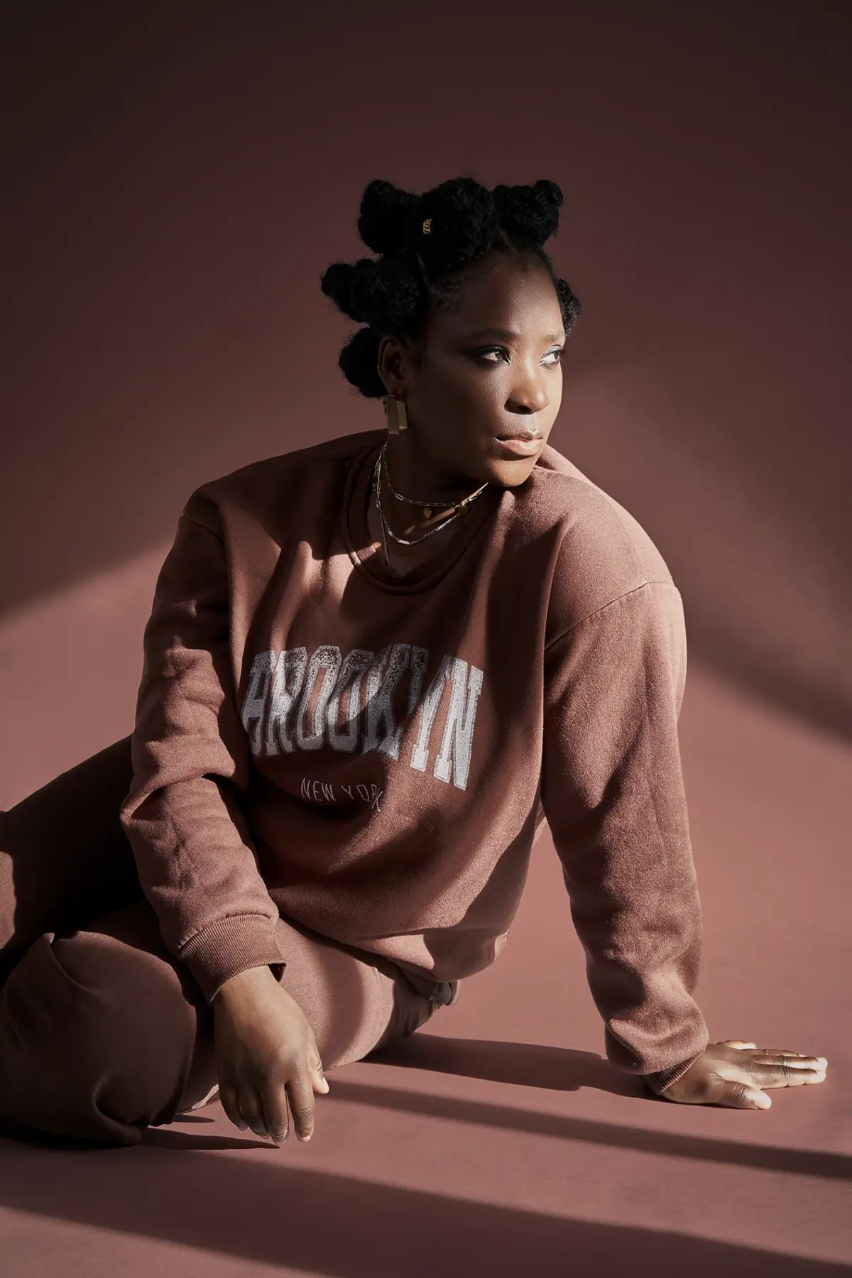 Studioaufnahme einer Frau im braunen Sweatshirt mit „Brooklyn“-Aufdruck; Bantu-Knot-Frisur, goldene Ketten, seitlicher Blick; weiches Licht und warmer Hintergrundton.
