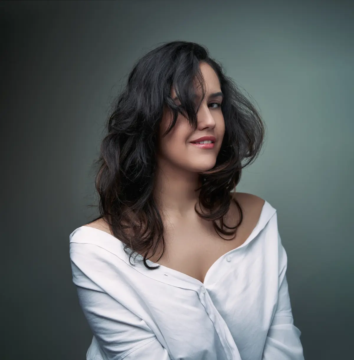 Frau mit dunklem, leicht zerzaustem Haar sitzt vor graugrünem Hintergrund; trägt ein weißes Hemd, das locker über die Schulter rutscht; lächelt dezent mit natürlicher Ausstrahlung.