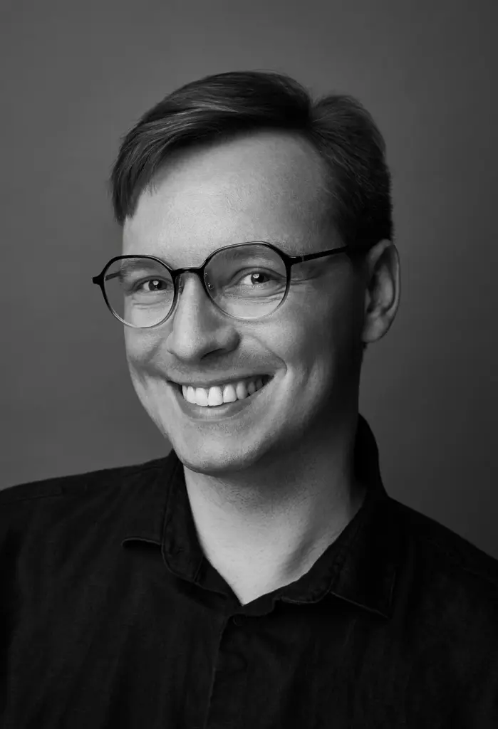 Lars Brodnicki ist frontal im Bild zu sehen. Er trägt eine runde Brille und hat kurzes Haar. Sein Gesicht ist freundlich und leicht lächelnd. Die Aufnahme ist in Schwarzweiß gehalten, wodurch die feinen Abstufungen von Licht und Schatten sehr deutlich wirken. Der Hintergrund ist hell und gleichmäßig, ohne Struktur, sodass der Fokus vollständig auf seinem Gesicht liegt. Die Lichtquelle kommt von vorne, wodurch sein Lächeln weich und harmonisch erscheint.