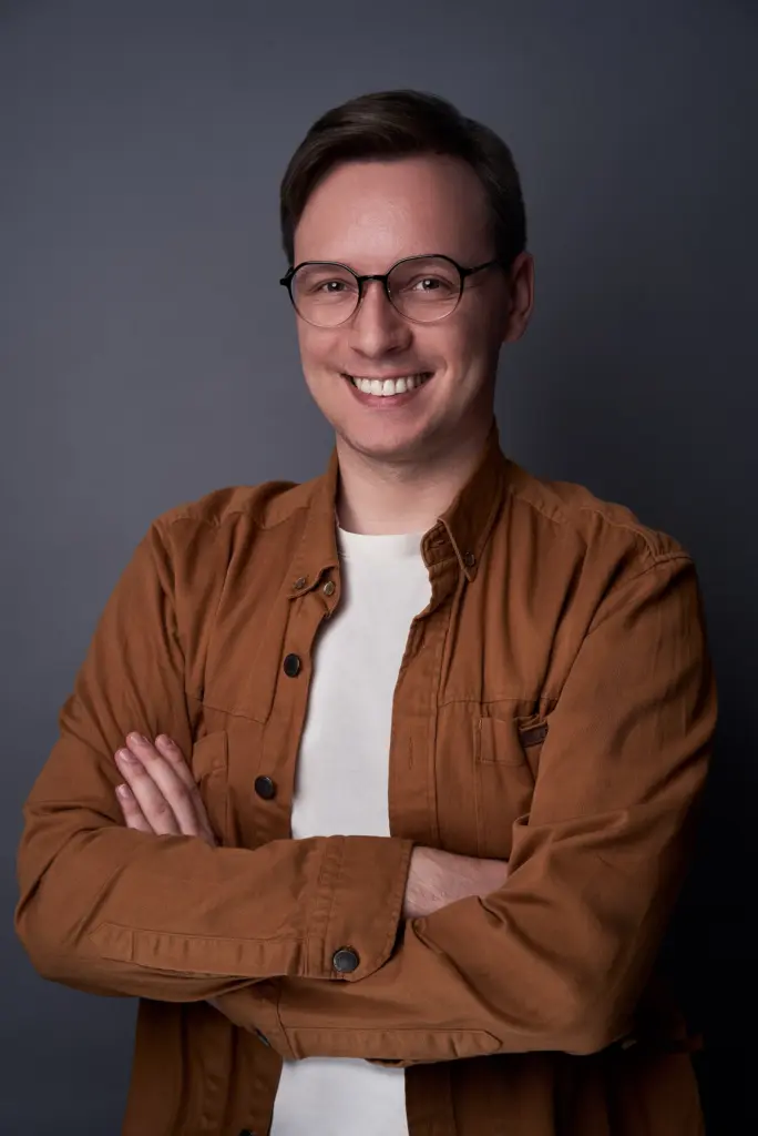 Lars Brodnicki steht vor einem dunklen, ruhigen Studiohintergrund. Er trägt eine dunkle Brille und darüber ein braunes Hemd, das offen über einem hellen T-Shirt liegt. Seine Arme sind locker vor der Brust verschränkt, was ihm eine entspannte, selbstbewusste Haltung verleiht. Sein Lächeln ist warm und offen. Das Licht fällt weich von vorne auf sein Gesicht und betont sanft die Konturen, ohne harte Schatten zu erzeugen.