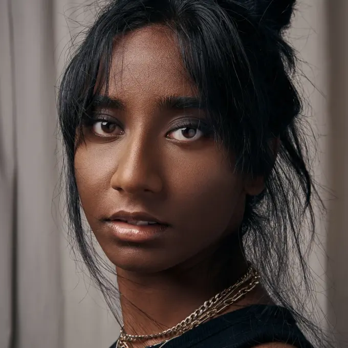 Nahporträt einer Frau mit lockerem Dutt und herausfallenden Strähnen; dezentes Make-up, glänzende Lippen, feine Ketten; neutraler Vorhanghintergrund, weiches frontales Licht.