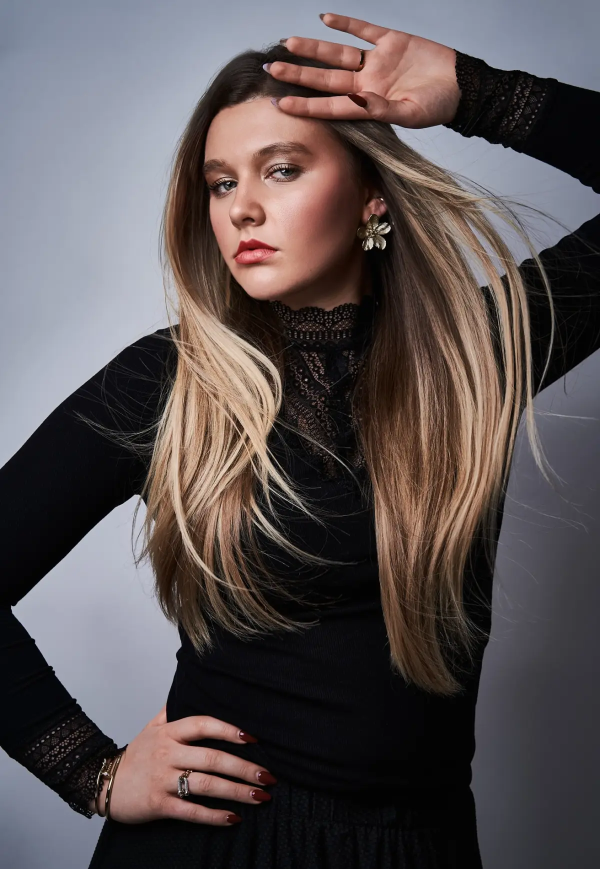 as Model steht vor einem neutralen, hellgrauen Studiohintergrund. Sie trägt ein schwarzes, langärmeliges Oberteil mit feiner Spitze am Kragen und an den Ärmelbündchen. Ihr Haar ist lang, glatt und in sanften, natürlichen Blond- und Brauntönen gehalten. Eine Hand liegt mit leicht gebeugtem Ellbogen auf ihrer Hüfte, während die andere erhoben ist und lässig nahe an ihrer Stirn ruht, wodurch eine dynamische Linienführung entsteht. Das Gesicht wirkt ruhig und selbstbewusst, der Blick ist leicht zur Seite gerichtet. Sie trägt auffällige, goldfarbene Blumenohrstecker sowie feine Armbänder und Ringe, die den eleganten Look ergänzen.