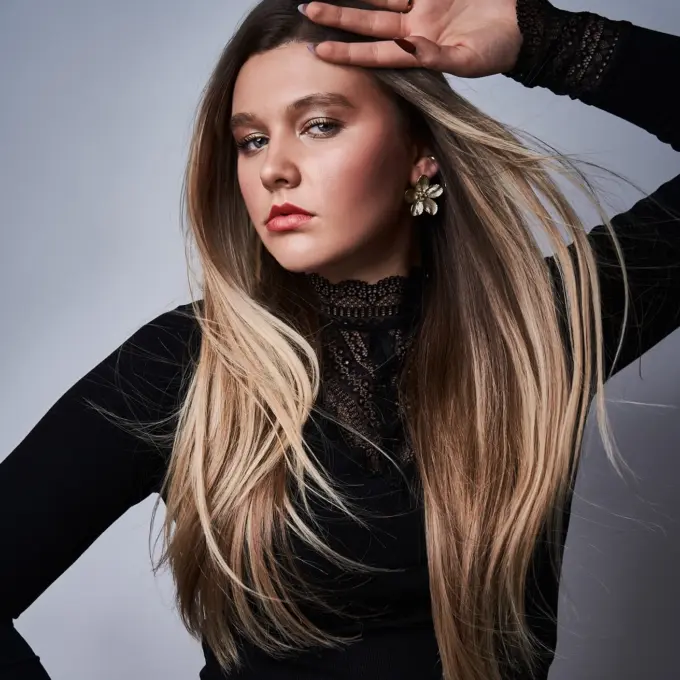 as Model steht vor einem neutralen, hellgrauen Studiohintergrund. Sie trägt ein schwarzes, langärmeliges Oberteil mit feiner Spitze am Kragen und an den Ärmelbündchen. Ihr Haar ist lang, glatt und in sanften, natürlichen Blond- und Brauntönen gehalten. Eine Hand liegt mit leicht gebeugtem Ellbogen auf ihrer Hüfte, während die andere erhoben ist und lässig nahe an ihrer Stirn ruht, wodurch eine dynamische Linienführung entsteht. Das Gesicht wirkt ruhig und selbstbewusst, der Blick ist leicht zur Seite gerichtet. Sie trägt auffällige, goldfarbene Blumenohrstecker sowie feine Armbänder und Ringe, die den eleganten Look ergänzen.